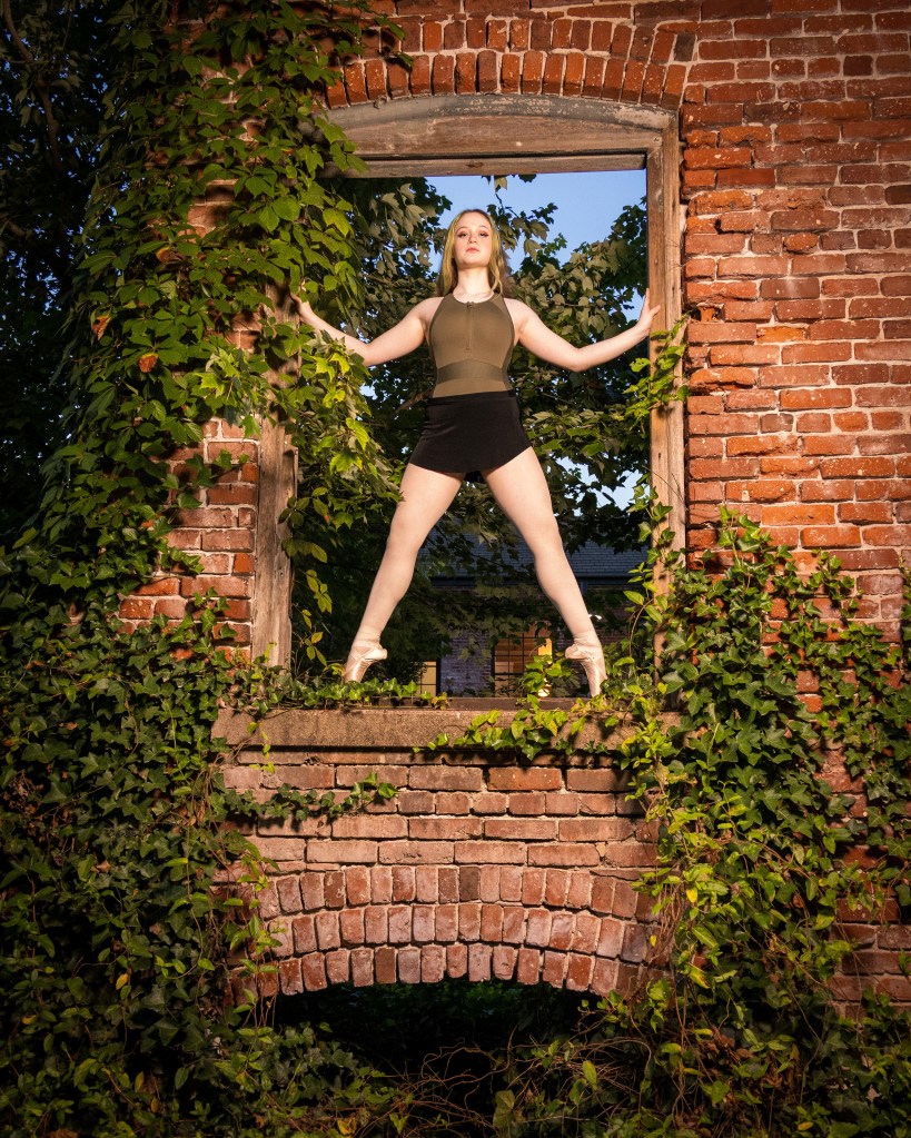 A dance ambassador in a black leotard performing a powerful pose with arms extended, standing in front of a weathered brick wall inside the historic Silk Mill.