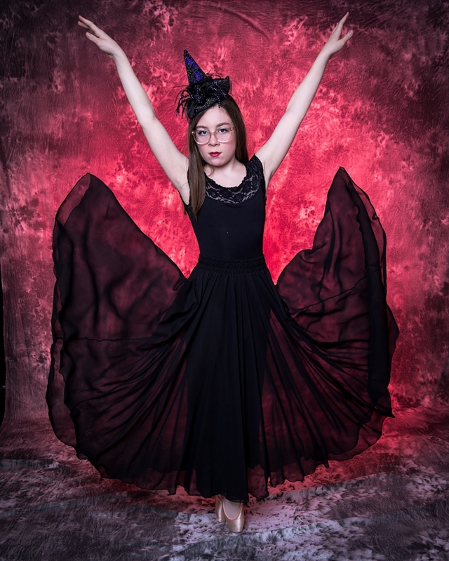Young dancer in a dramatic black gothic-style gown and a witch's hat, posing against a dark background with vibrant purple smoke effects.