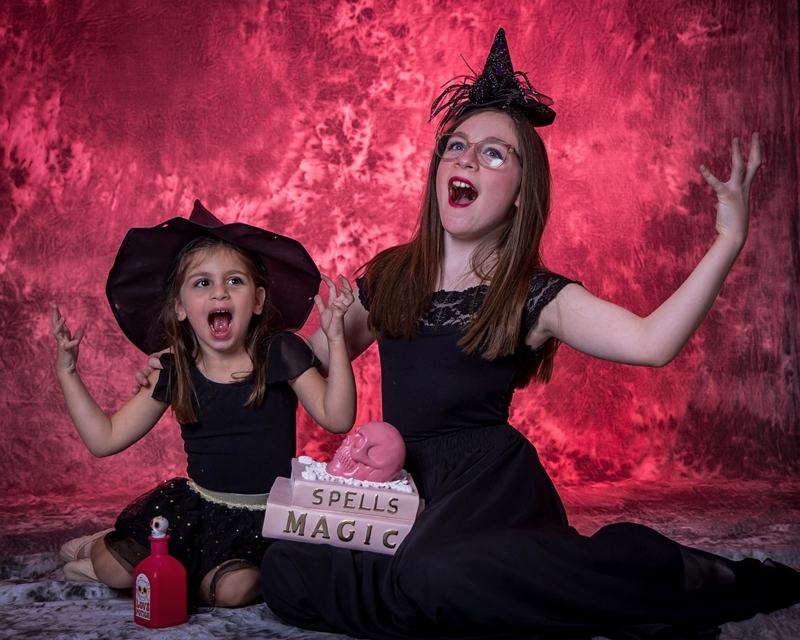 Young dancer in a dramatic black gothic-style gown and a witch's hat, posing against a dark background with vibrant purple smoke effects.