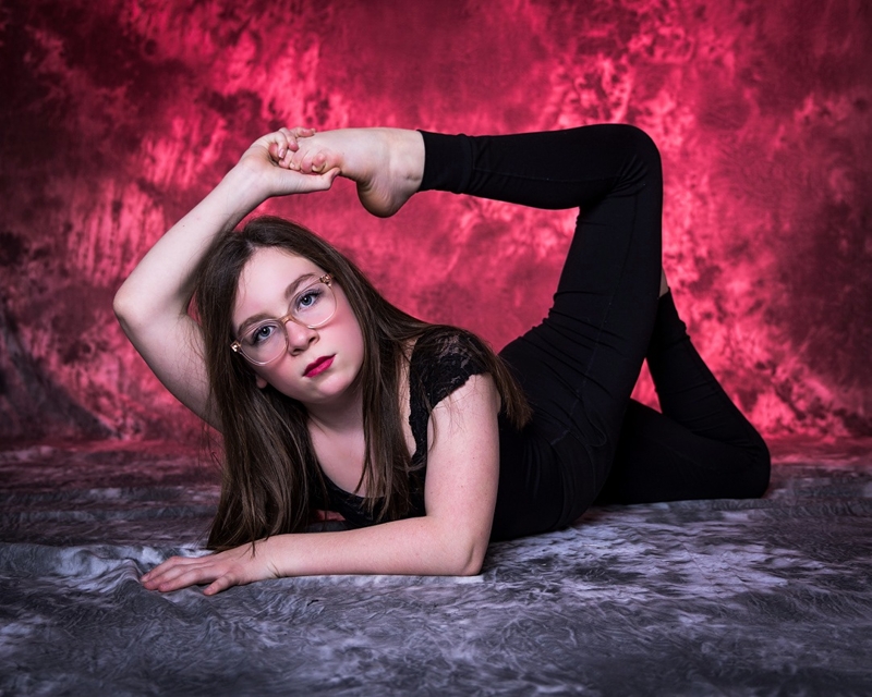 Young dancer in a dramatic black gothic-style gown and a witch's hat, posing against a dark background with vibrant purple smoke effects.