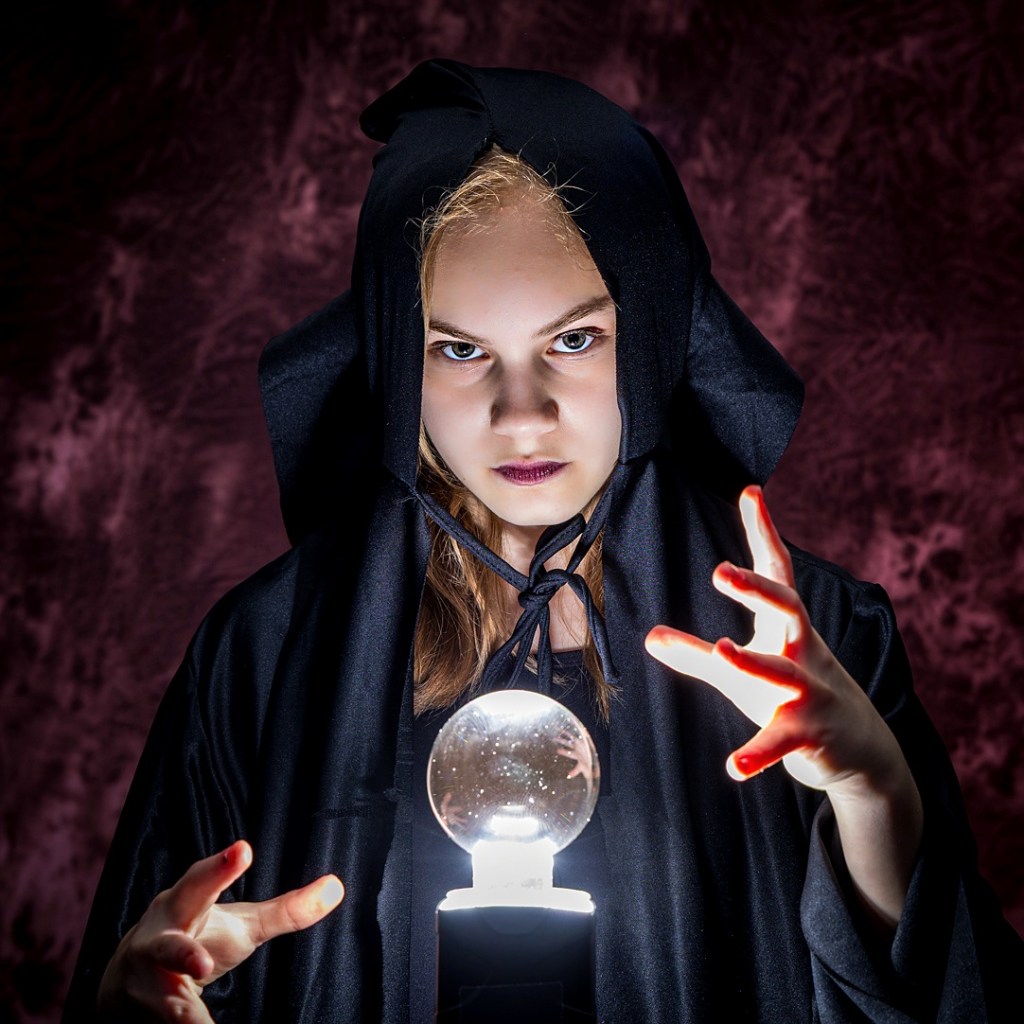A young dancer, Sydney, wearing a black hooded cloak, looking intensely at the camera. She is holding her hands over a glowing crystal ball on a pedestal, with a dramatic smoky purple and black textured background.