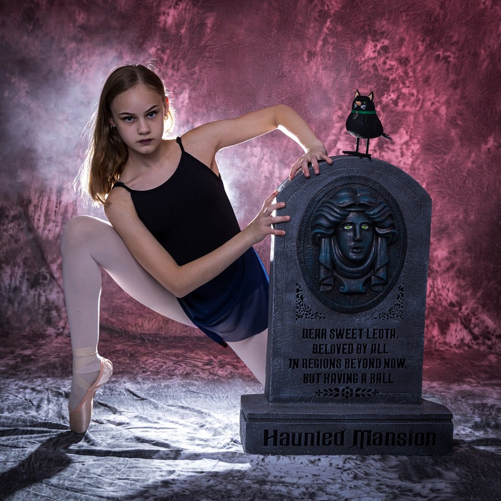 A young dancer, Sydney, in a black leotard and white pointe shoes, posing next to a "Haunted Mansion" tombstone. She has a serious expression and is set against a textured magenta and smoky background with a black cat prop atop the gravestone.