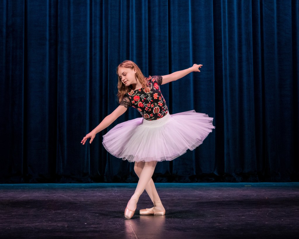 A young female ballet dancer in a white professional pancake tutu with a black floral embroidered bodice. She is posing on a dark stage in front of deep blue velvet curtains, smiling with one arm extended and her feet in a ballet position.