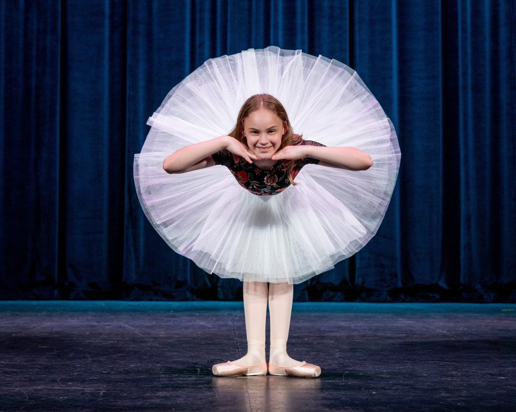 A young female ballet dancer in a wide white professional tutu and black bodice, smiling and posing with her hands on her chin. She is centered on a dark stage in front of closed blue velvet curtains.