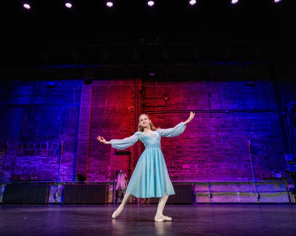 ance Ambassador Sydney performing on a stage with vibrant purple and blue brick-wall backdrop lighting. She is wearing a light blue long-sleeved dance dress and pointe shoes, captured in an elegant open-arm pose.