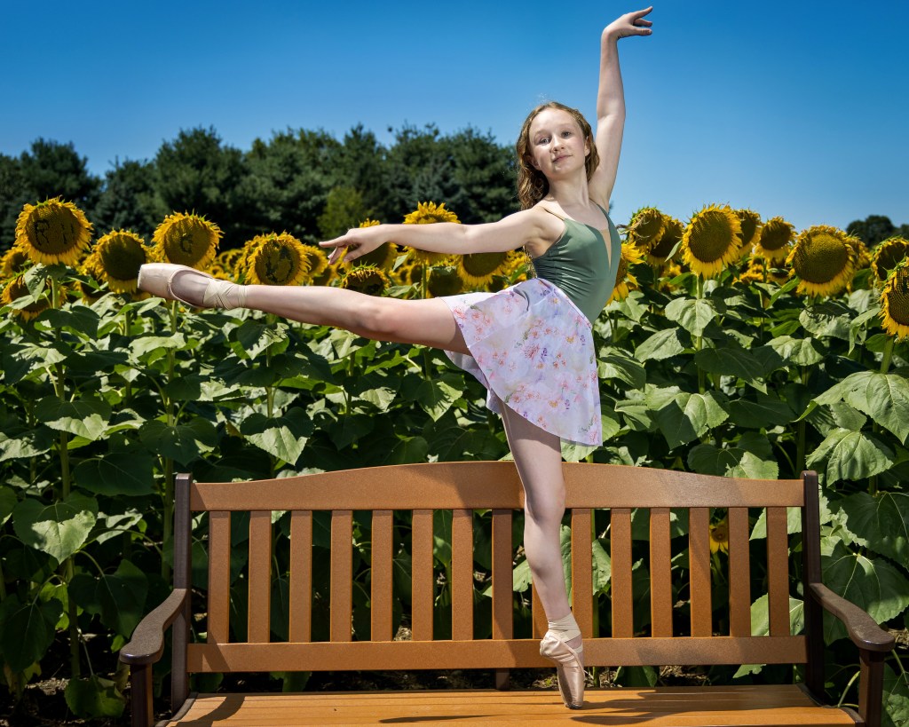 A young ballet dancer, Lily, performing a graceful pose on a wooden bench in a vibrant sunflower field. She is wearing a light green leotard and white pointe shoes, with one leg extended and an arm raised, surrounded by tall yellow sunflowers under a clear blue sky.