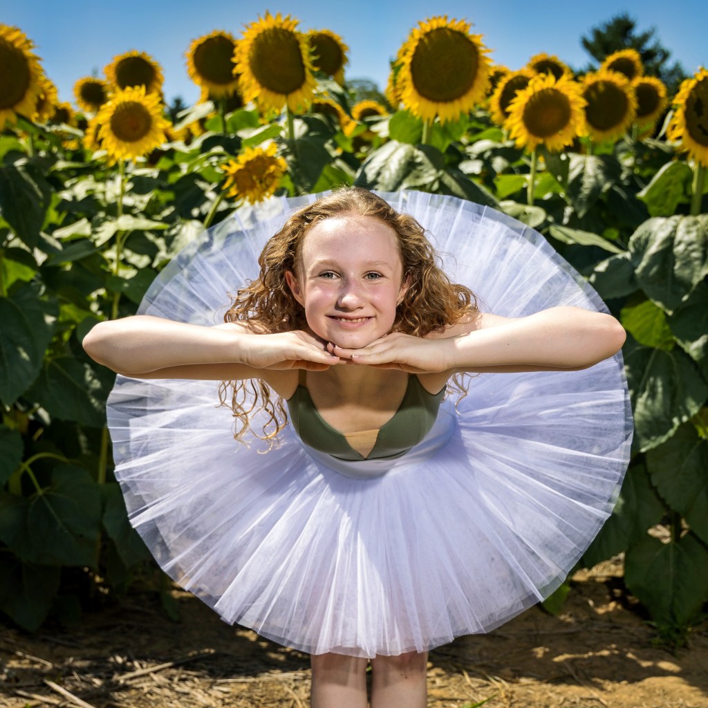 Ballet dancer Lily performing a standing pose among tall sunflowers. She is on pointe with her arms in an elegant fifth position, wearing a green leotard that complements the natural greenery of the sunflower field under a bright sky.