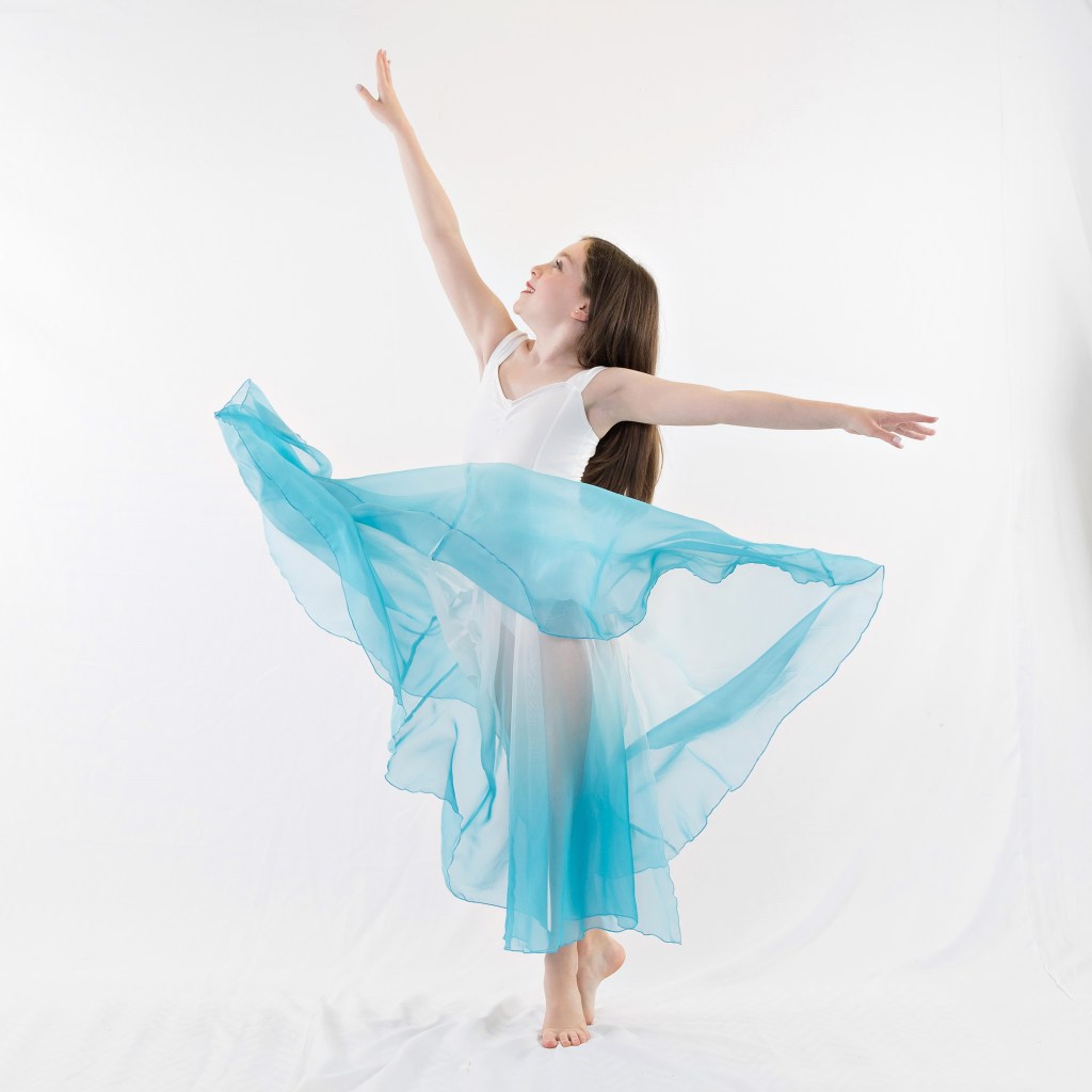 Young ballerina in a light blue and white tutu performing a graceful pose with one arm extended, set against a soft blue and white painterly studio backdrop.