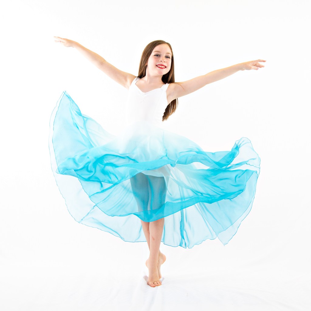 Young ballerina in a light blue and white tutu performing a graceful pose with one arm extended, set against a soft blue and white painterly studio backdrop.