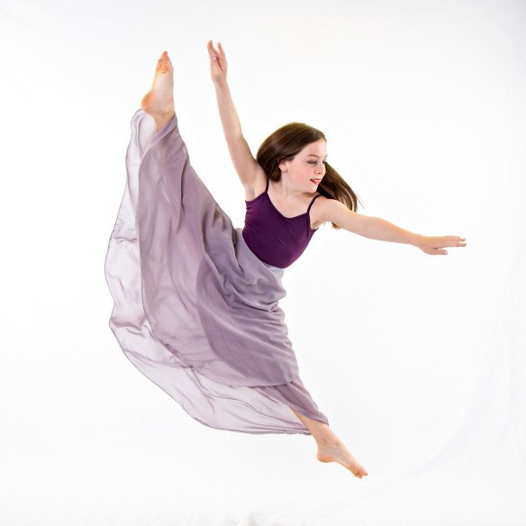 Young ballerina Olivia Klein wearing a purple leotard and a long, flowing sheer white skirt, performing a dynamic dance pose in a studio.