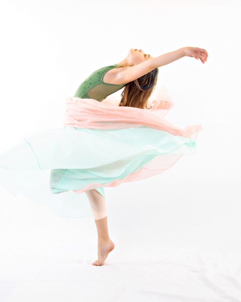 A female dancer, Sydney, in a green leotard performing a dynamic pose. She is surrounded by layers of flowing, translucent white and mint green fabric that creates a sense of motion against a bright white background.