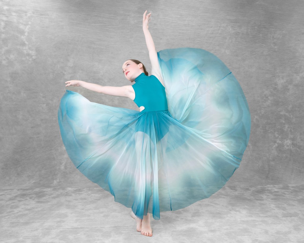 Young ballerina in a light blue and white tutu performing a graceful pose with one arm extended, set against a soft blue and white painterly studio backdrop.