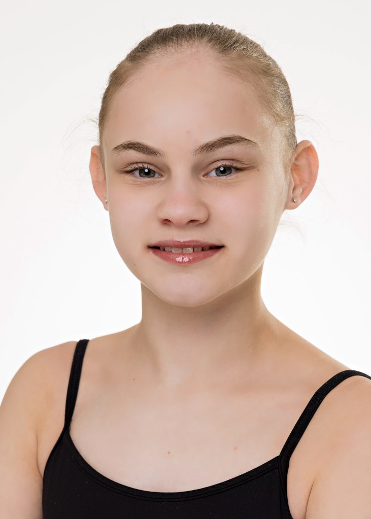 A professional headshot of a young female dancer with her hair in a tight bun, wearing a black spaghetti-strap leotard, smiling against a plain white studio background.