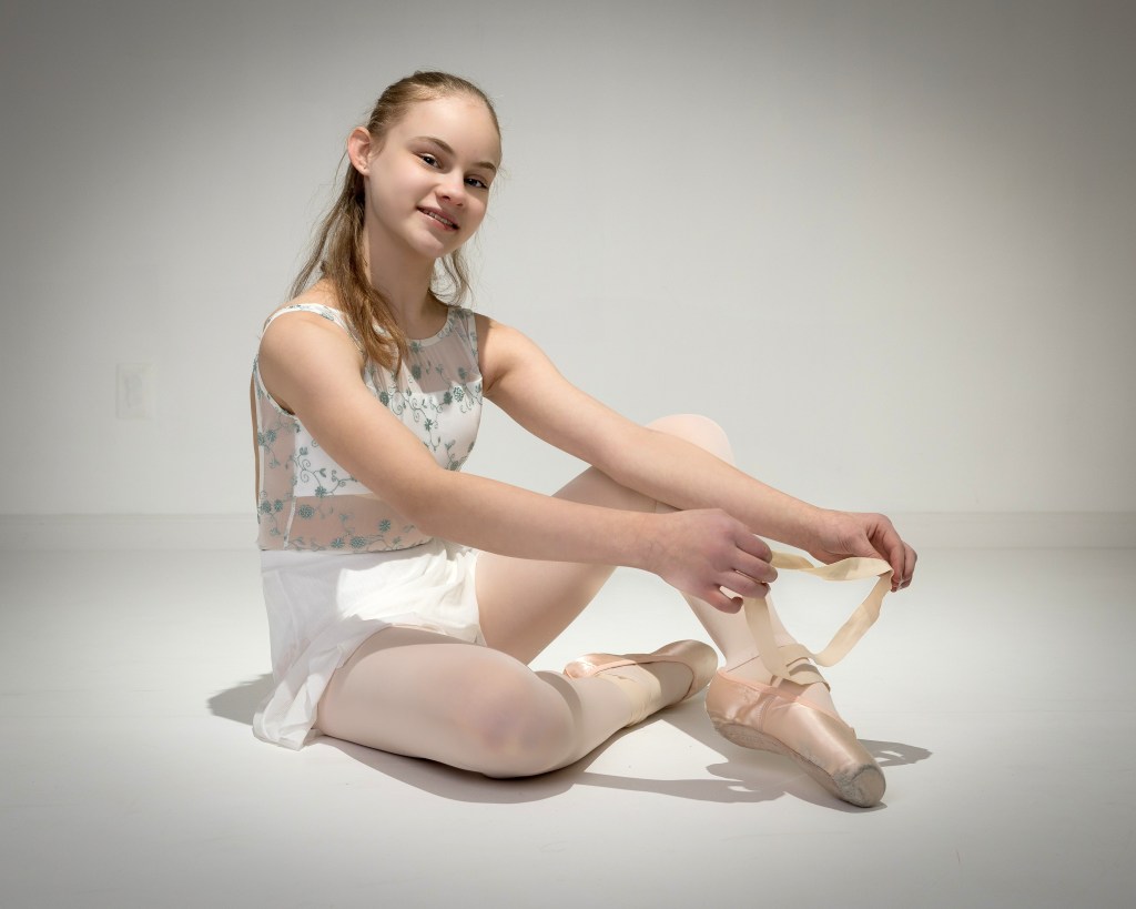 A young ballet dancer, Zoey, sitting on a studio floor and smiling at the camera while tying the ribbons of her pointe shoes. She is wearing a white floral leotard and a white skirt against a light grey background.