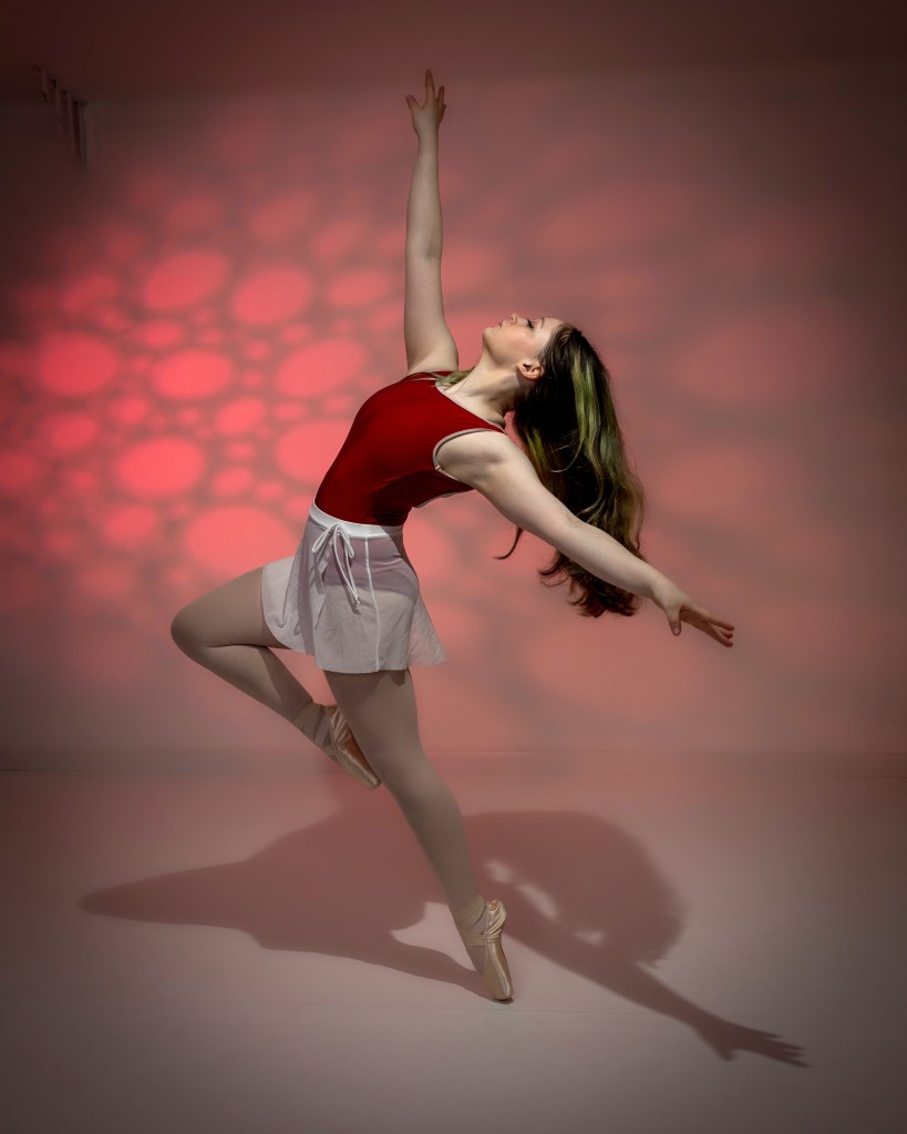 A creative dance portrait of Zoey in a green leotard and white tutu, posing elegantly on one pointe shoe against a vibrant purple and pink "Valentine" studio backdrop.