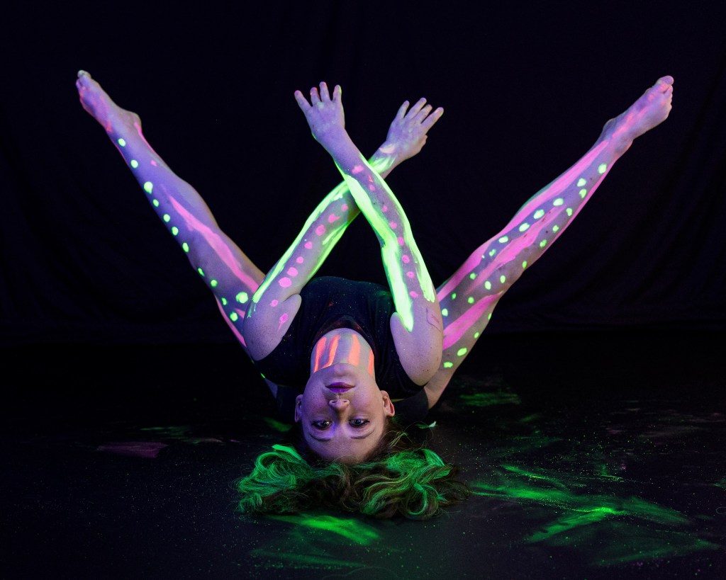 A dancer in a black leotard with neon green and purple body paint posing with arms forming a 'W' shape under UV blacklight.