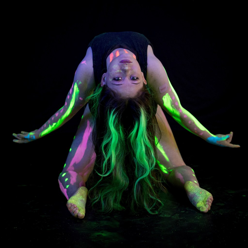 Close-up portrait of a dancer with vibrant neon green and purple face paint and glowing makeup, illuminated by UV studio lighting.