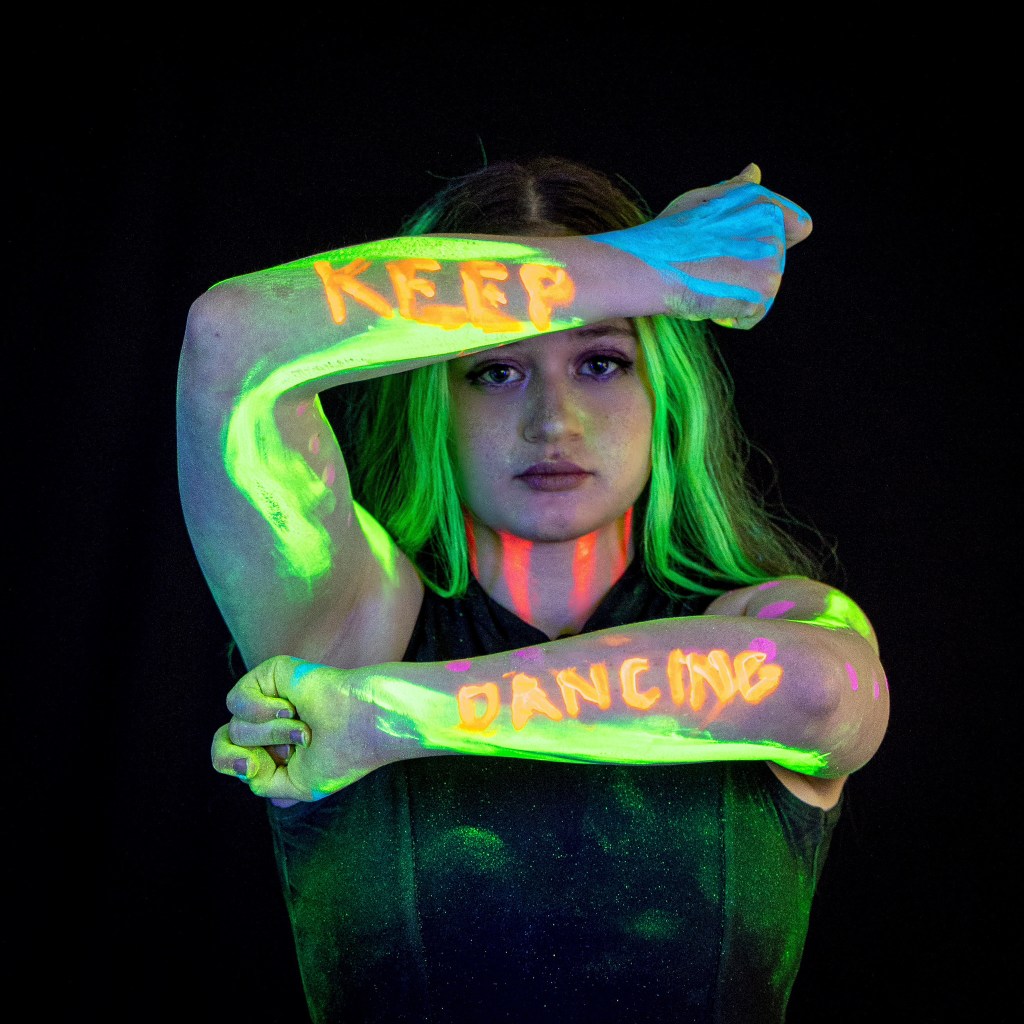 A dancer wearing a glowing 'DANCING' headband and neon body paint, holding a neon prop against a dark studio background.